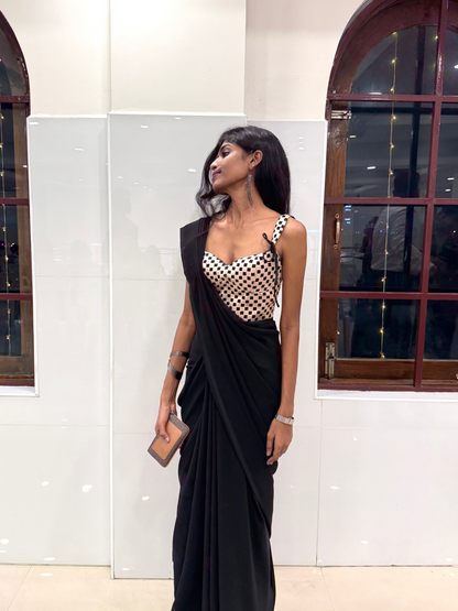 Woman in a black saree with a patterned blouse standing in front of a white wall with arched windows.